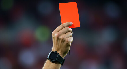 A close-up of a referee's hand holding up a red card on a soccer field. This image directly symbolizes punishment, expulsion, or serious foul play.