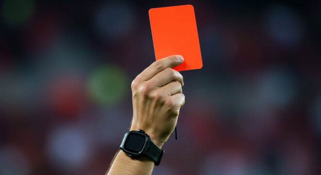 A close-up of a referee's hand holding up a red card on a soccer field. This image directly symbolizes punishment, expulsion, or serious foul play.