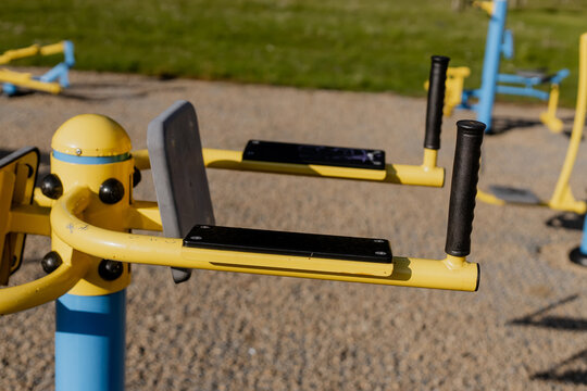 Close-up of outdoor dip station for upper body training in public fitness park, yellow and blue metal exercise equipment on gravel surface in sunlight