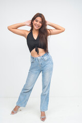 Cheerful young Asian woman in black top and denim pants posing casually with hands raised behind head and a smile. Isolated on a white background.