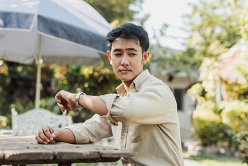 An annoyed young Asian man checks his watch outside, appearing frustrated and impatient while waiting for someone to arrive.