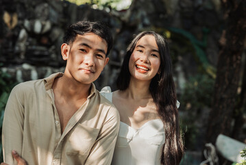 A cheerful young Southeast Asian man stands beside his smiling girlfriend under the shade of a tree enjoying the peaceful afternoon together.