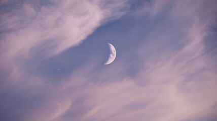 Crescent moon partially obscured by wispy clouds during twilight, creating a serene and ethereal atmosphere with soft pastel colors