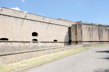 A female tourist stands facing the massive stone walls of the Forte Spagnolo in L'Aquila, Italy, on a sunny day.