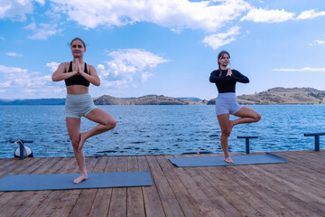 Women practicing yoga on a wooden dock by the lake during a sunny day