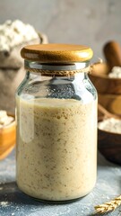 Glass jar of creamy sourdough starter, surrounded by flour and baking tools