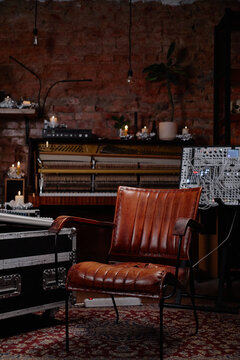 Empty vintage leather chair standing in front of open upright piano and modular synthesizer in music studio with exposed brick wall and candles