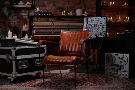 Empty vintage leather chair standing in music studio surrounded by analog audio equipment, modular synthesizer, reel to reel tape recorder, candles, exposed brick wall background