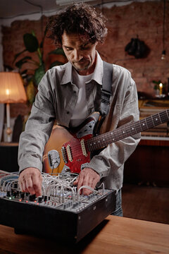 Caucasian man playing electric guitar while adjusting modular synthesizer, standing indoors with focused expression, engaging in music production or sound design