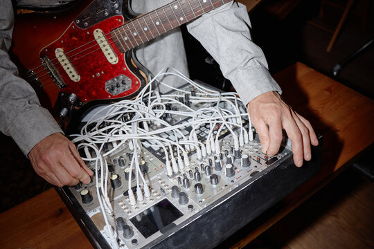 Caucasian man adjusting modular synthesizer and playing electric guitar, hands visible operating audio equipment on wooden table, creating electronic music setup