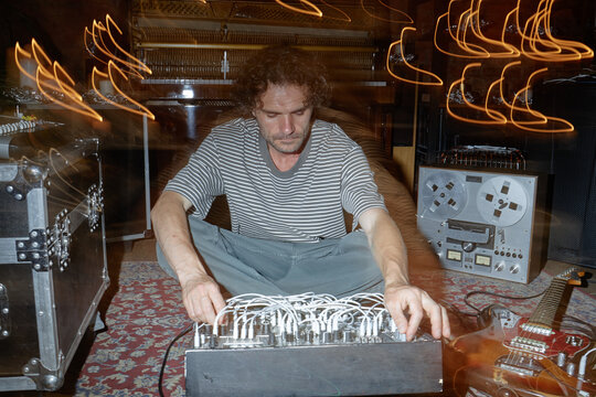 Caucasian man sitting on floor operating modular synthesizer surrounded by vintage audio equipment and electric guitar in music studio with blurred light trails