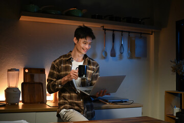 Young man sits in the kitchen at the bar counter works in laptop and drinks a coffee. Home atmosphere, work in the evening after a hard day. Home office.