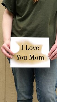 A Woman&rsquo;s Hands Holding Up a Gold Painted Stencil That Says I Love You Mom in front of a Tan Backdrop