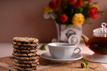 elegant tea setting with oatmeal chocolate cookies, white cup, glass teapot, and vibrant flowers on vintage newspaper surface in soft light, style, comfort, leisure, relaxing, gourmet, layered