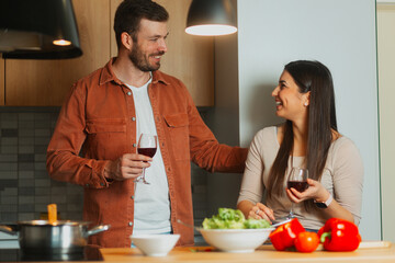 Couple enjoying time together in the kitchen, preparing fresh food and testing red wine. Domestic lifestyle with love, cooking, and shared moments at home