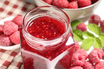Delicious raspberry jam in glass jar, fresh berries, mint and checkered napkin on white table, closeup