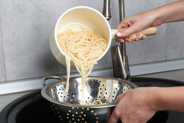 Woman draining boiled tagliatelle pasta into colander at sink in kitchen, closeup