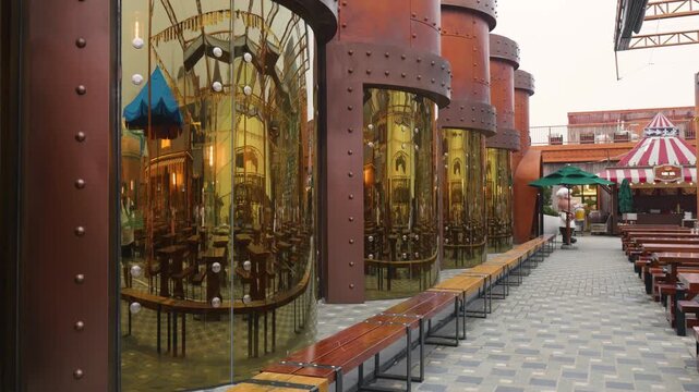 Golden cylindrical mirrors with rivets reflecting a restaurant interior, wooden benches lining the walkway, and a chef statue standing near a food stall with a striped awning at ba na hills