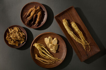 Simple flat lay of ginseng roots in a rectangular tray, paired with supporting herbal ingredients in round bowls on black background. Minimal and elegant look.