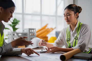 Two female architects wearing safety vests are examining blueprints and discussing project details in a brightly lit office, with a scale model of a building visible in the background