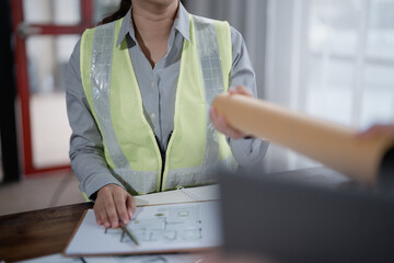 Engineer wearing a safety vest handing over blueprints to a colleague while pointing at a house plan on a clipboard during a collaborative meeting focused on project development