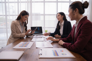 Three asian businesswomen sitting at wooden table analyzing financial charts and discussing marketing strategy using smartphone and laptop in modern office