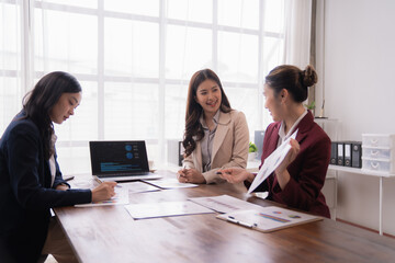 Three Asian businesswomen collaborating at a wooden table in a bright office, discussing marketing strategies while analyzing charts and graphs on paper and laptop