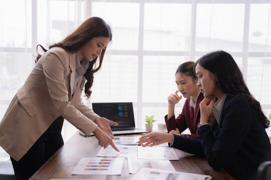 Three Asian businesswomen analyzing financial charts and engaging in discussions about their project during a meeting in a modern office, showcasing teamwork and strategic planning