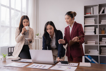 Three asian businesswomen are having a meeting using laptop and analyzing financial charts, graphs and paperwork while drinking coffee in modern office