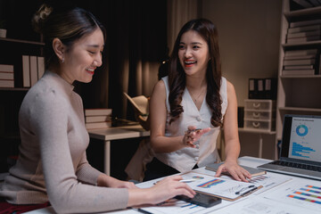 Fototapeta premium Two smiling Asian businesswomen are discussing a marketing strategy using charts, graphs, and a laptop, working late at night in the office to achieve their business goals