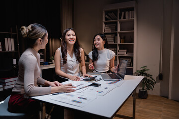 Three Asian businesswomen smiling and collaborating late at night in the office, working together on a laptop while analyzing documents and discussing project details