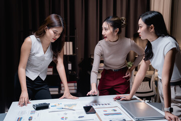 Three asian businesswomen leaning on a desk and arguing about negative financial results showing on some documents during a late night meeting in the office