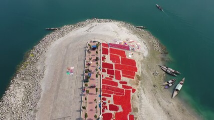 Bogura, Bangladesh - 21 August 2025: Aerial view of bright red chilies spread out to dry on a sandy peninsula surrounded by boats and calm turquoise water.