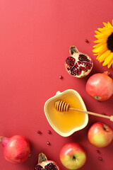 Flat lay of apples, pomegranates, sunflower, and honey in yellow apple-shaped dish with dipper on red background. Festive Jewish New Year still life