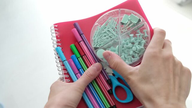 A person is holding a red notebook with a green section in the middle. The notebook is filled with colorful pens and scissors