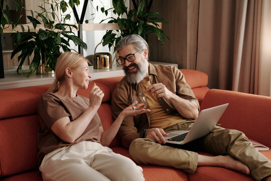 Mature man and woman in love enjoying spending time together at home sitting on sofa having snack and talking while watching film online on laptop