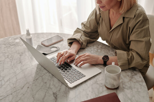 High angle view shot of mature Caucasian woman spending morning at home sitting at table typing something on laptop