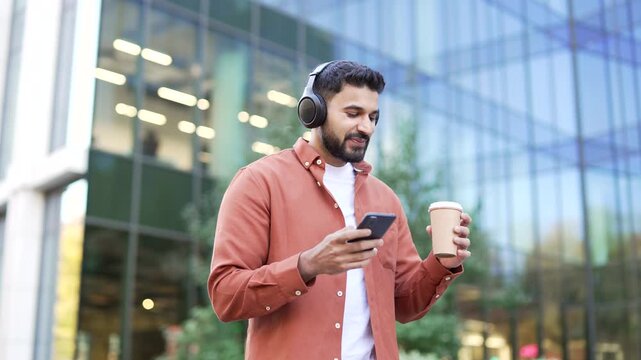 Happy businessman in wireless headphones walking on street looking at phone outside modern office building Smiling confident handsome bearded man with cup of coffee in hand enjoying listening to music