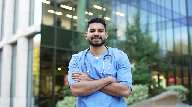Portrait of a smiling doctor in a blue coat looking at the camera, standing outside near the hospital building. Happy handsome male medical worker physician posing on the street with crossed arms
