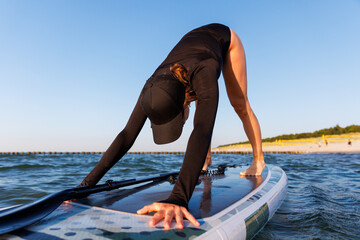 Young adult slim woman perform Yoga Downward-Facing Dog surfing SUP paddleboard Baltic Sea waves warm sunset sun lights summer day. Sunlight sea ocean waves enhance harmony body, strength mindfulness