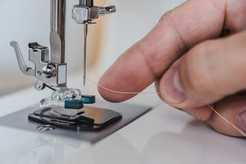 Male fingers putting thread in needle loop on electric modern sewing machine for start sewing. Manually threading sewing machine needle with fingers, close-up of precise process