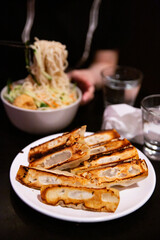 Crispy pan fried dumplings served on white plate in restaurant setting, background shows bowl of noodles being lifted with chopsticks