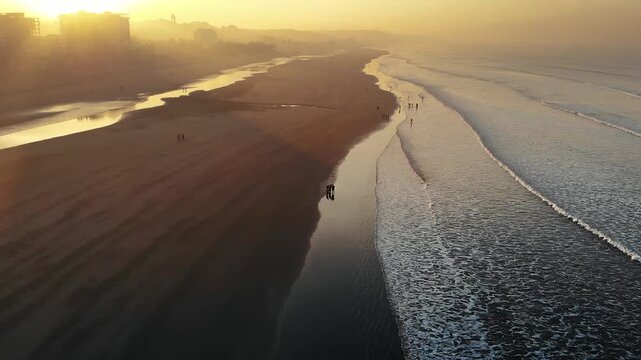 Cox's Bazar, Bangladesh - 20 August 2025: Aerial view of a vast beach where people stroll along the water's edge as the sun casts a golden glow over the scene.