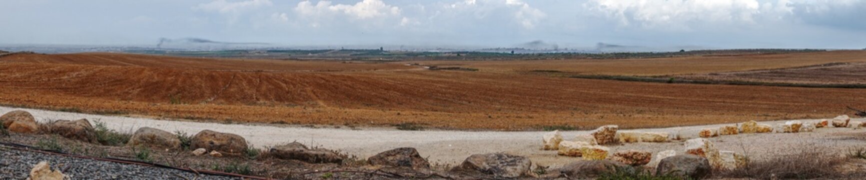 View of rich brown fields under a vast sky, contrasted by a rough white path and scattered rocks, with the horizon fading into a distant city, Mefalsim, South District, Israel.