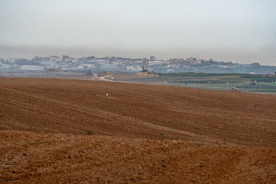 View of a freshly tilled field under a soft, diffused light, leading towards a distant cityscape shrouded in a gentle haze, Mefalsim, South District, Israel.