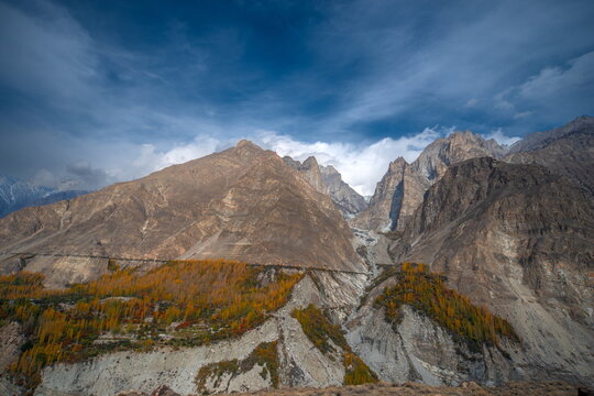 View of rugged, towering mountains with a stripe of autumnal trees against a backdrop of a dramatic sky, Hunza Nagar, Gilgit Baltistan, Pakistan. - Powered by Adobe