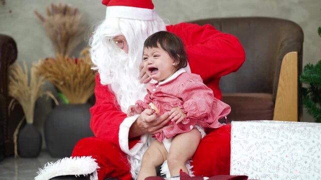 A santa claus carrying and comforting crying baby girl in red dress on armchair at home on Christmas holiday