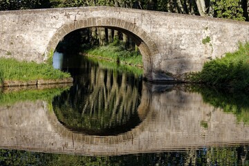 Fototapeta premium Stone arch bridge reflects in a calm river surrounded by lush greenery, creating a symmetrical view.
