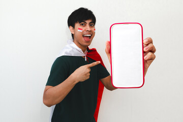 Excited Indonesian man holding a flag and pointing at smartphone with happy expression on white isolated background, perfect for independence celebration and advertisement.