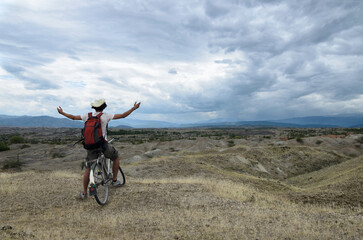 Traveler on a bycicle embracing the sky with his hands up in the prairie